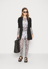 Blazer preto sobre uma blusa ajustada com padrão botânico e leggings em branco, cinza e preto. Segura uma bolsa preta acolchoada e usa sandálias de plataforma pretas.