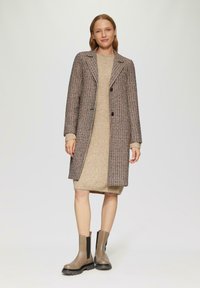 Houndstooth jas in bruin en crème, enkel gebuttonneerd met twee knopen. Beige gebreide jurk en bruin enkelschoenen met zwarte elastische zijden.