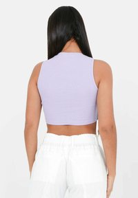 Žena s dlouhými černými vlasy, oblečená v bezramínkovém levandulovém crop topu a bílých kalhotách s vysokým pasem, zobrazená zezadu.