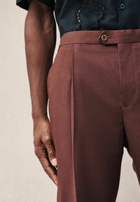 Brown tailored trousers with a smooth texture and several front pleats, featuring a button closure at the waist. Dark shirt with embroidered detail.