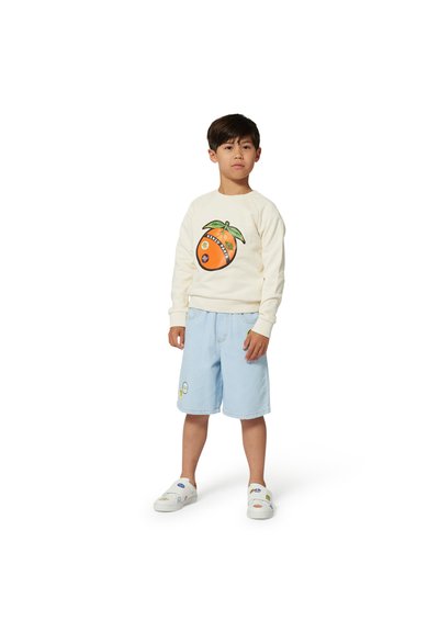 Sweatshirt crème avec un motif graphique orange et un design de feuilles vertes, associé à un short en denim bleu clair avec des accents en patch et des chaussures blanches à enfiler.