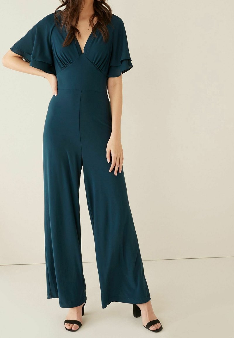 Femme portant une combinaison pantalon large bleu sarcelle foncé avec des manches volantées et un décolleté en V profond, associée à des talons carrés noirs ouverts.