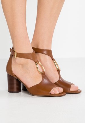 Tacones abiertos marrones de cuero con tiras en el tobillo y detalles de hebillas doradas, usados en los pies sobre un fondo blanco.