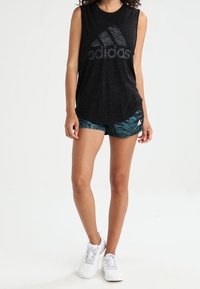 Musta ilmanhihainen urheilupaita, jossa on teksturoitu pinta ja harmaa adidas-logo, yhdistettynä vihreisiin kuosiin shortsien ja valkoisten lenkkareiden kanssa.