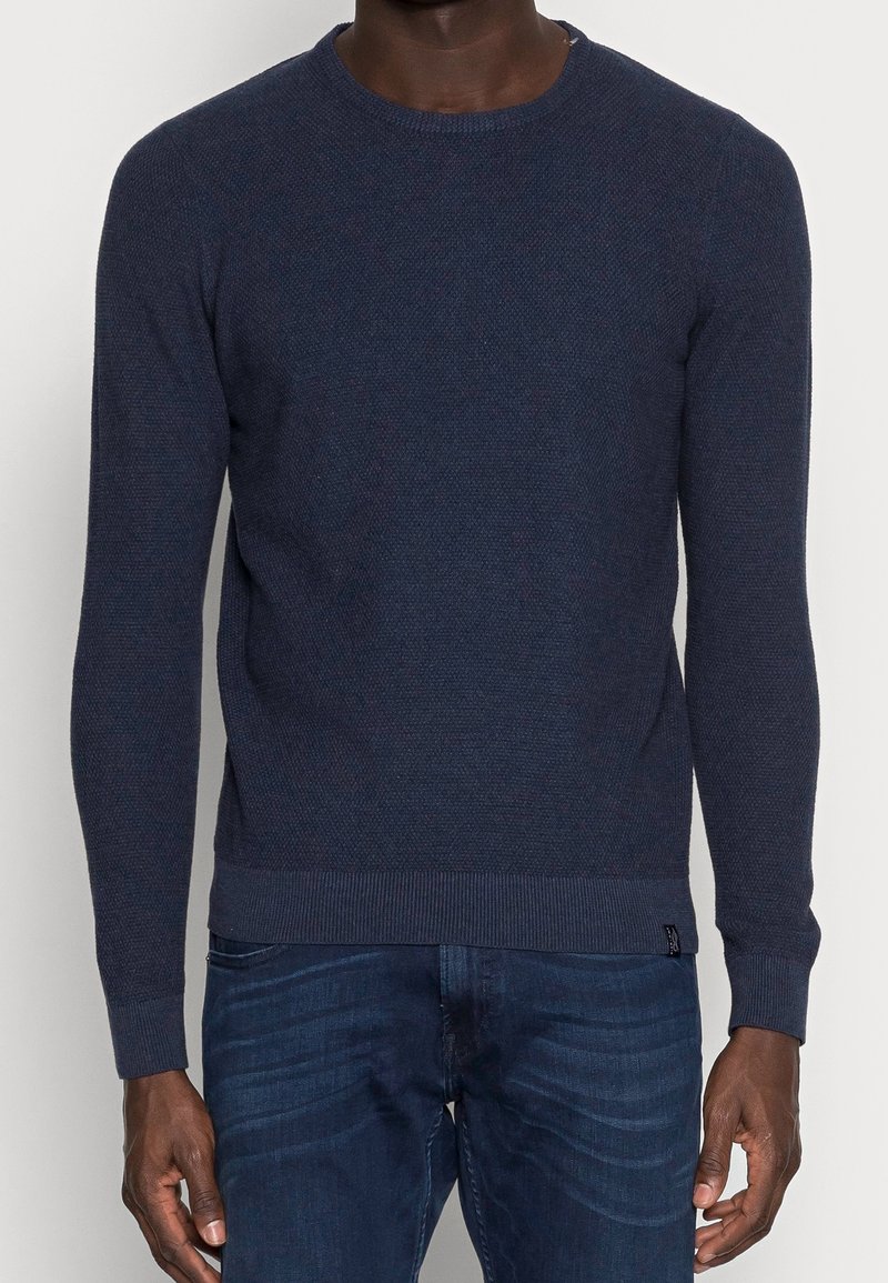 Homme portant un pull à manches longues texturé bleu foncé et un jean bleu de lavage moyen, debout les bras détendus le long du corps.