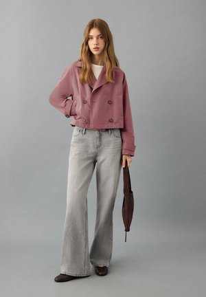 Oblečena svetlo roza dvoredna jakna do pasu z dvema velikima gumboma, kombinirana z širokimi sivo-jeans hlačami z zvončastimi hlačnicami in temno rjavimi čevlji.