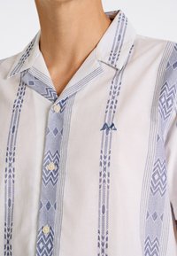 Camisa de manga corta en tejido ligero con patrones geométricos azules, base blanca y cierre de botones. Presenta un pequeño logo bordado.