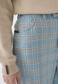 Blauwe geruite broek van een gestructureerde stof, met lichtblauwe, bruine en witte lijnen, en een zijzak met een bruine label.