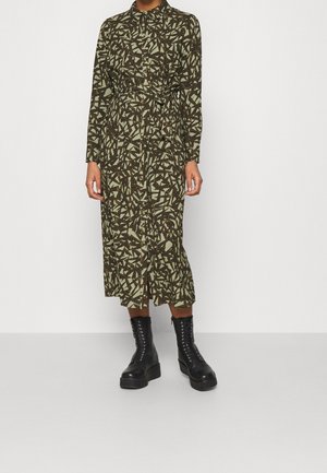 Osoba ubrana w midi sukienkę na długi rękaw z guzikami, z abstrakcyjnym czarno-beżowym wzorem oraz czarne platformowe botki na sznurowadła.