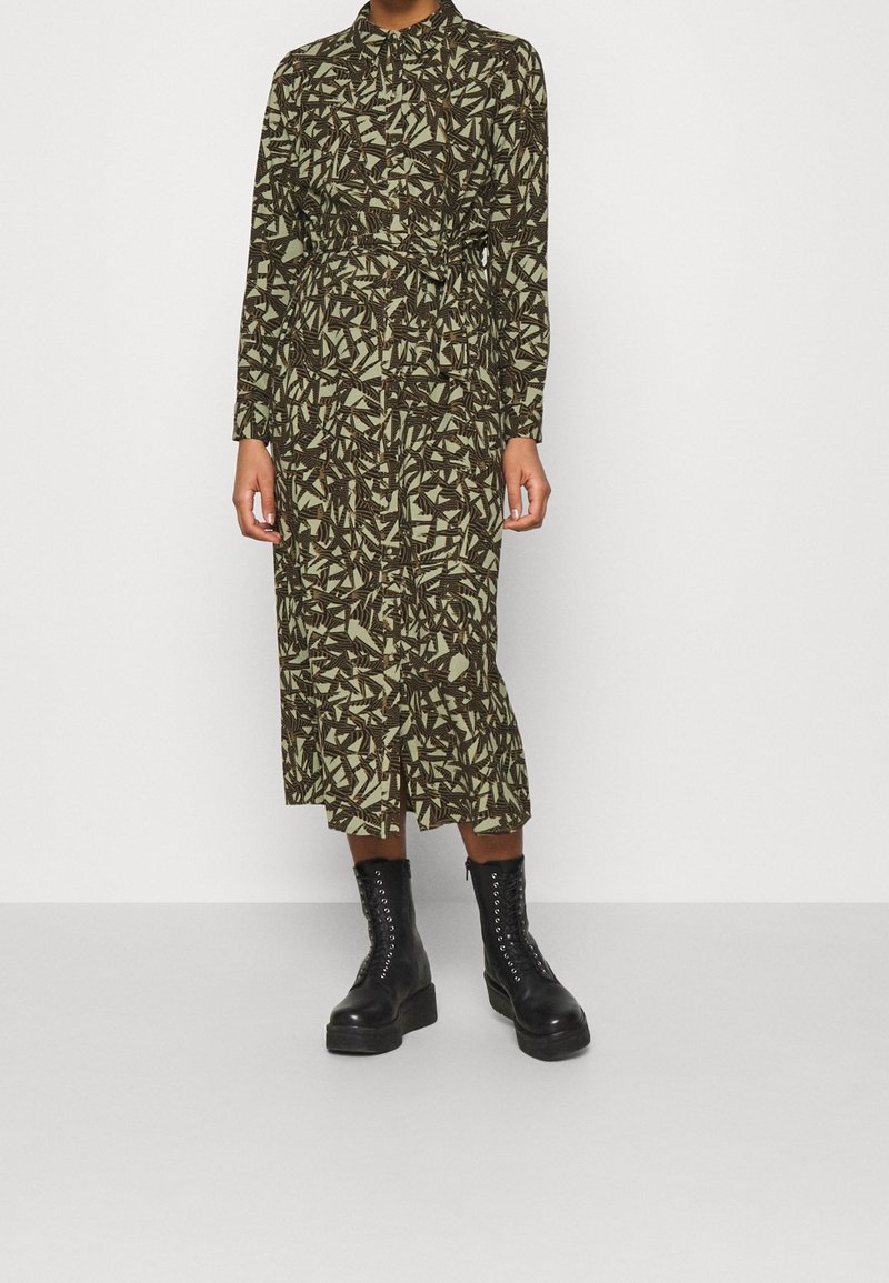 Personne portant une robe midi à manches longues boutonnée avec un motif abstrait noir et beige, et des bottes plateformes noires à lacets.