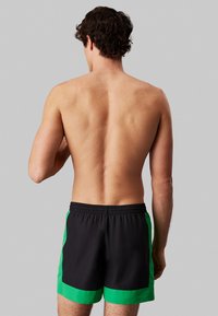 Czarne kąpielówki z zielonymi bocznymi panelami, elastyczny pas, długość powyżej kolan, lekki materiał, gładka tekstura.