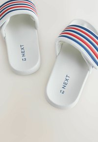 Sandalele albe tip papuci au o talpă texturată, o bretea superioară lată cu dungi albastre, roșii și albe și inscripționarea în relief a mărcii.