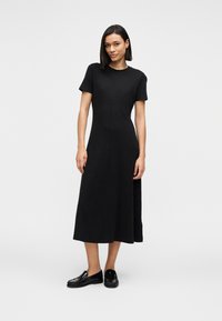 Μαύρο μίντι φόρεμα με κοντά μανίκια, στρογγυλή λαιμόκοψη, ριμπ υφή και ευθεία σιλουέτα, συνδυασμένο με γυαλιστερά μαύρα μοκασίνια.