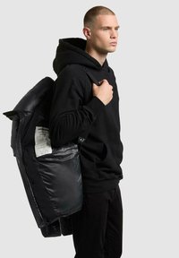 Zwarte gewatteerde jas met glanzende textuur, voorzien van een label aan de binnenkant, gedragen over een zwarte hoodie. De pose van het model benadrukt het design van de jas.