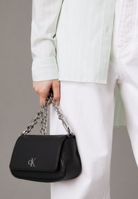 Borsa a tracolla in pelle nera con catena argentata, dotata di pattina e logo impresso, tenuta in mano accanto a una camicia a righe verde chiaro.