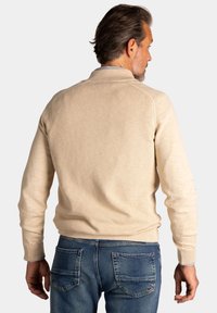 Lichtbeige gebreide sweater met een gestructureerde afwerking, voorzien van een hoge kraag en ribboorden, gecombineerd met blauwe denim jeans.