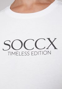 Camiseta blanca de algodón con un atrevido texto negro que dice "SOCCX" y "EDICIÓN TIMELESS", con cuello redondo y mangas cortas. Textura suave.