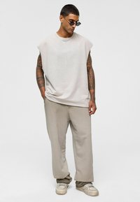Lichtgrijze oversized mouwloze shirt met een ronde hals, gecombineerd met losse beige broeken. Draagt witte sneakers en donkere zonnebril.