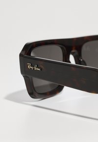 Ochelari de soare Ray-Ban din băț de țestoasă, cu ramă rectangulară, lentile închise la culoare și detalii aurii cu logo-ul pe tempă. Textură netedă din plastic.