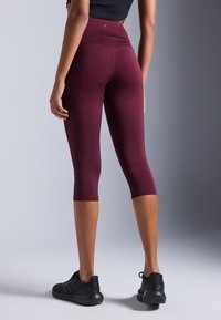 Maroon capri tajice s visokim strukom, glatka tkanina, prilagodljiv dizajn i kontrastni šavovi; u kombinaciji s crnim sportskim cipelama.
