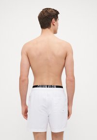 Bijele kupaće gaće s elastičnom trakom u struku s crnim logotipom "CALVIN KLEIN". Sadrže jedan stražnji džep i imaju glatku teksturu.