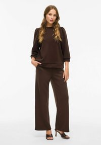 Brun genser med puffede ermer, kombinert med vide brune bukser. Glatt tekstur, avslappet design og lang passform.