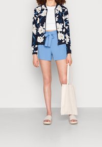 Floral bomber jacket in navy and white, white crop top, light blue shorts with tie, beige slip-on sandals, and a beige tote bag.