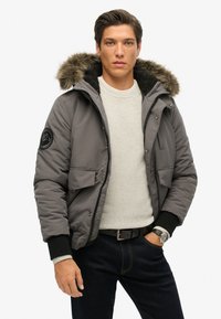 Giacca isolante grigia con cappuccio foderato in pelliccia, zip, bottoni a pressione, due tasche frontali e polsini a coste sopra un maglione bianco strutturato.