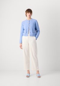 Hellblau gestrickte Strickjacke mit rundem Ausschnitt, goldenen Knöpfen und Vordertaschen, kombiniert mit weißen weit geschnittenen Hosen und hellblauen Riemchensandalen.