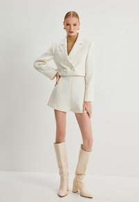 Witte tweed blazer met dubbele knopen, gecombineerd met bijpassende shorts. Model draagt kniehoge beige blokhaklaarzen en een bruine coltrui.