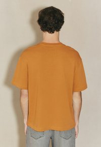 Kurzärmliges, Rundhals-T-Shirt aus orangefarbenem Baumwollstoff. Hat eine lockere Passform und eine glatte Textur. Rückansicht mit schlichter Gestaltung.