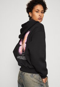 Mlada osoba s kovrčavom kosom koja nosi naušnice u obliku obruča, crnu hoodicu s velikim ružičastim leptirićem i tekstom na leđima, u kombinaciji s svijetloplavim trapericama.