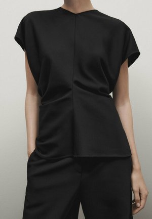 Woman wearing loose black sleeveless top with a seam detail down the center paired with black pants, hands relaxed by sides.