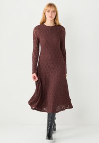 Brązowa koronkowa sukienka z długimi rękawami, teksturowanym wzorem i rozkloszowanym dołem. Noszona z czarnymi kozakami do kolan.