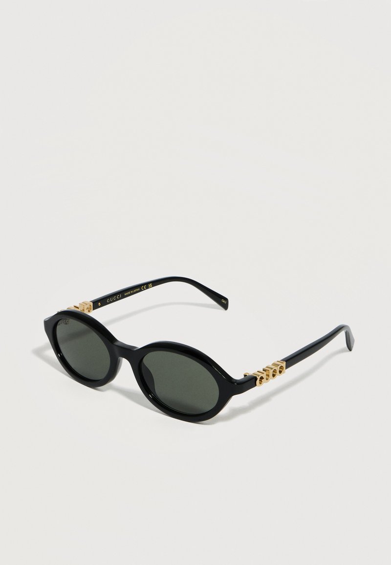 Μαύρα οβάλ γυαλιά ηλίου Gucci με σκουρόχρωμους φακούς και χρυσές λεπτομέρειες με το λογότυπο στα πλαϊνά, τοποθετημένα σε φωτεινή επιφάνεια.