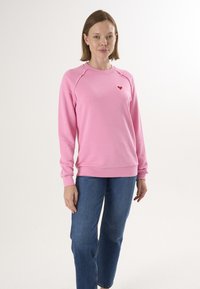Felpa rosa con scollo rotondo, maniche raglan e un piccolo cuore rosso sul petto. Abbinata a jeans blu a gamba dritta.