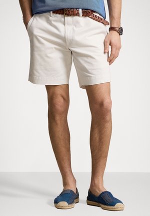 Homme portant un short beige avec une ceinture tressée marron, des chaussures bleues sans lacets et une chemise bleue, debout les mains dans les poches.