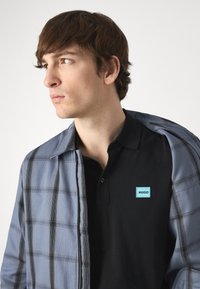Herremote herre med en blå rutete jakke med glidelås og svart poloskjorte. Poloskjorten har en liten blå "HUGO"-lapp på brystet.