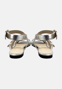 Un paio di sandali piatti argento con cinturini alla caviglia e schiena aperta, mostrati da dietro su uno sfondo bianco.