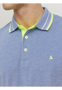 Niebieska koszulka polo z fakturowanej tkaniny, zielonym kołnierzykiem w białe paski i trzema guzikami. Mały akcent w postaci logo na klatce piersiowej.