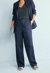Un costume en lin bleu marine avec un blazer ajusté et un pantalon à jambes larges. L'ensemble comprend un top en col en V, une montre en argent et un sac à main bleu clair.
