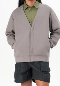 Šedá mikina s výstřihem do V a dlouhými rukávy, jednobarevná struktura, boční kapsy, nošena přes zelenou košili s límečkem a spojena s černými šortkami.