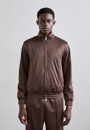 Brun zip-up jakke med høy krage og elastiske mansjetter. Laget av glatt stoff, med en logo på venstre bryst og glidelåslommer.