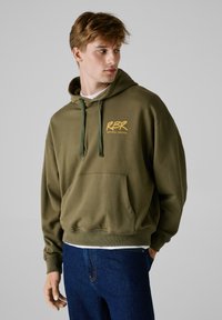 Haut à capuche vert olive en tissu doux, avec une poche avant, une capuche avec cordon de serrage et un logo "RBR" jaune sur la poitrine. Associé à un jean bleu.