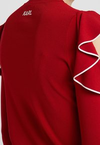 Maglione rosso con trama a coste, maniche arricciate con bordi bianchi e una delicata ricamo "KARL" sul retro.