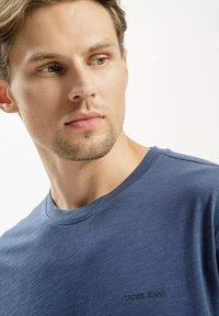 Granatowa koszulka typu T-shirt z okrągłym dekoltem, wykonana z teksturowanej tkaniny z subtelnym przeszyciem. Posiada logo "CROSS.JEANS" na dolnym lewym boku przodu.