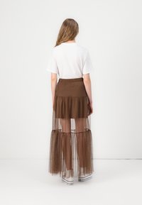 Falda marrón con volantes y superposición de tul transparente, cinturilla elástica y camiseta blanca. Llevada con zapatillas blancas para crear contraste.