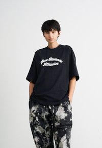 Une personne se tient les mains dans les poches, portant un t-shirt noir "New Balance Athletics" et un pantalon en tie-dye noir et blanc devant un fond simple.