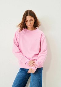 Kvinna med axellångt brunt hår, klädd i en rosa sweatshirt och blå jeans, som justerar sin ärm mot en enfärgad vit bakgrund.