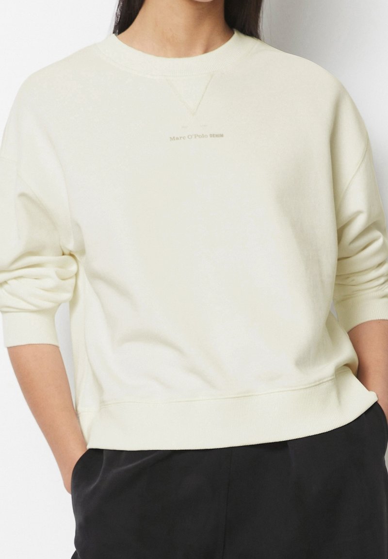 Sudadera beige claro con cuello redondo y puños acanalados, presenta un logo sutil en el pecho, confeccionada con un tejido suave y texturizado.